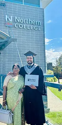 A graduate in cap and gown standing with a family member outside their college on graduation day