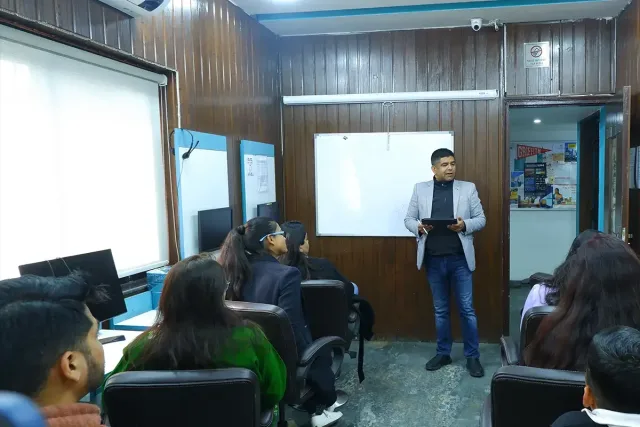 A trainer teaching from the whiteboard while students listen from the front rows, with computer workstations along the wall behind.
