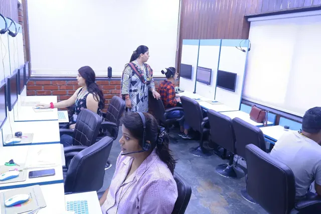 Students at computer workstations with headsets on while a trainer walks between the desks.